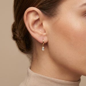 Könnte beinhalten: Ein goldener Creolen-Ohrring mit einem rechteckigen, klaren Edelstein-Anhänger. Der Ohrring wird am Ohr einer Person mit heller Haut und braunem Haar getragen. Das Design ist schlicht und elegant.