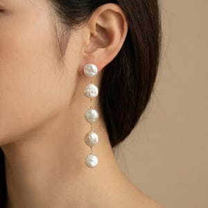 May include: A pair of elegant dangle earrings. Each earring features five large, lustrous white pearls suspended from delicate gold-coloured chains. The earrings are shown against a neutral background, highlighting their sophisticated design. A classic jewelry piece.