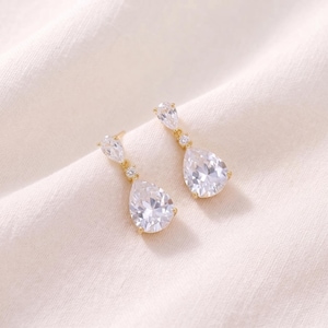 May include: A pair of gold-tone drop earrings featuring a small round clear stone at the top and a larger pear-shaped clear stone suspended below. The metal setting is gold-coloured, and the stones are faceted to reflect light with a clear, brilliant appearance.