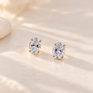 May include: A pair of stud earrings featuring oval-cut clear stones set in gold-toned metal. Each earring is secured with four prongs, showcasing a faceted surface that reflects light, creating a brilliant appearance for these jewellery pieces.
