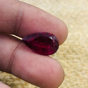 Può includere: Una gemma rosso scuro a forma di pera con una superficie sfaccettata. La pietra presenta una qualità traslucida e una finitura lucida, riflettendo la luce attraverso le sue molteplici superfici angolari, mostrando un ricco colore cremisi saturo.