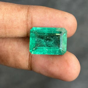 Puede incluir: Una esmeralda rectangular de tono verde vibrante con un corte facetado. La piedra muestra inclusiones internas visibles y vetas naturales en toda su estructura, exhibiendo una calidad translúcida con bordes afilados y bien definidos.
