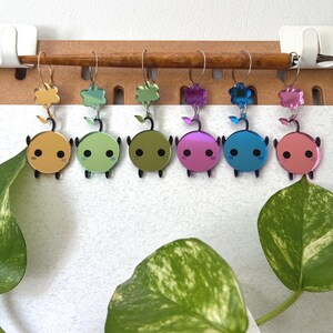 May include: A collection of seven colorful, mirrored acrylic earrings. Each earring features a round body with a face, small legs, and a leaf-shaped top. The earrings are in shades of gold, green, olive, pink, blue, and purple.