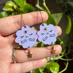 May include: A pair of lavender cloud-shaped earrings featuring a sleeping face with pink cheeks, a small gold star at the top, and a dangling gold star charm at the bottom. The earrings are attached to thin gold-toned metal hoop earring wires.