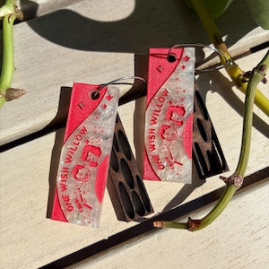 May include: A pair of rectangular dangle earrings featuring a red textured base and a translucent, glittery resin overlay. The resin displays the text ONE WISH WILLOW, small star icons, and two engraved faces. Each earring hangs from a silver hoop.