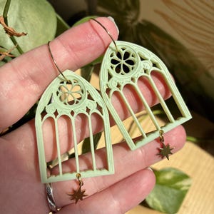 Puede incluir: Pendientes verde pálido con un diseño de ventana gótica. Los pendientes tienen aros dorados y pequeños colgantes de estrella. Los pendientes están sostenidos por la mano de una persona.