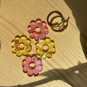 Puede incluir: Pendientes con forma de flor en tonos rosa y amarillo, con detalles dorados. También hay un par de pendientes de aro dorados. Los pendientes están sobre una superficie de madera.