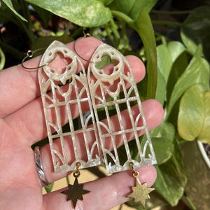 Puede incluir: Un par de pendientes con un diseño de ventana de catedral. Los pendientes son de color blanquecino con un patrón de rejilla y un dije de estrella dorada colgando de la parte inferior. Los pendientes se sostienen con aros dorados.