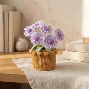 May include: A handcrafted crochet flower arrangement featuring lavender daisy-like blooms with purple centers and green leaves. The flowers are in a tan crochet pot with a scalloped edge, creating a charming, decorative piece.