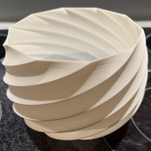 Peut inclure: Un pot de fleurs en forme de spirale de couleur crème avec une surface texturée. Le design présente une série de crêtes incurvées qui créent un effet tourbillonnant. Le pot est probablement fait d'un matériau durable.