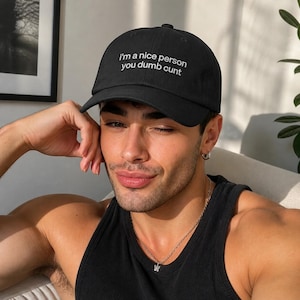 May include: Black baseball cap with the text "I'm a nice person you dumb cunt" in white. The cap is worn by a person wearing a black tank top and a silver necklace.