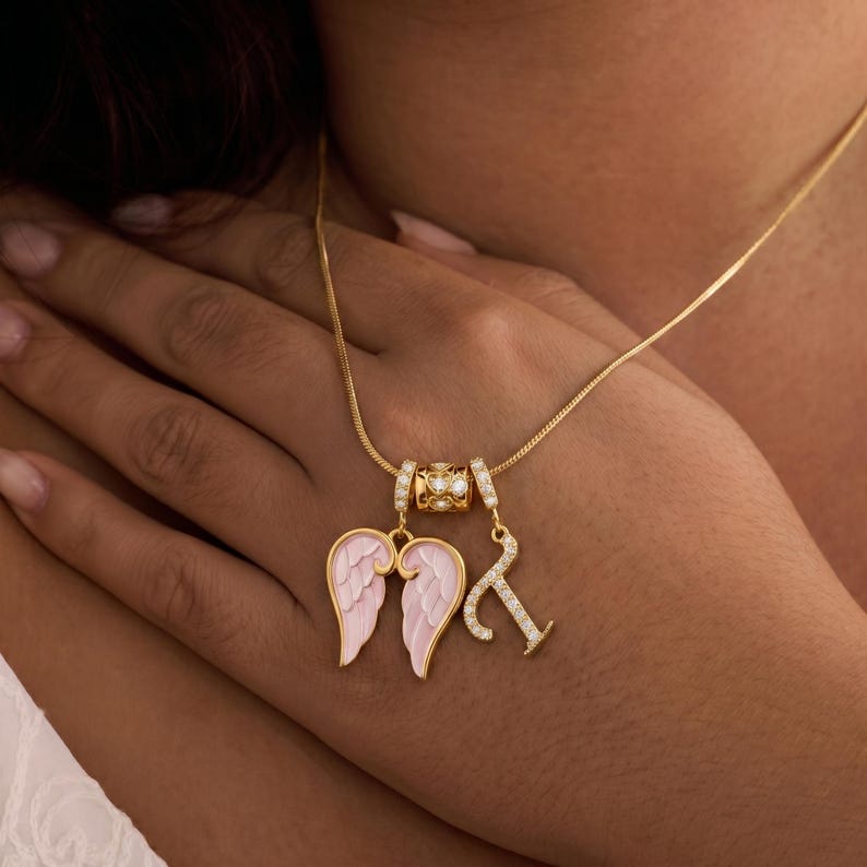 Puede incluir: Collar dorado con un colgante con alas de &aacute;ngel rosas y un charm con la letra "K", ambos adornados con detalles brillantes. El collar tiene una cadena delicada y una cuenta decorativa con piedras transparentes.