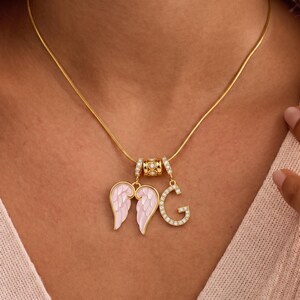 Puede incluir: Un collar dorado con un colgante que presenta alas de &aacute;ngel rosas y una letra "G" incrustada con peque&ntilde;os cristales transparentes. El collar tambi&eacute;n incluye una cuenta cil&iacute;ndrica decorativa con detalles de cristal.