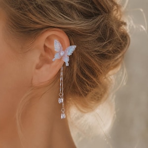 May include: An ear cuff featuring a translucent, iridescent light blue butterfly with a central crystal accent. Two delicate silver-toned chains hang from the butterfly, each ending in a small, clear, round faceted crystal charm that dangles below.