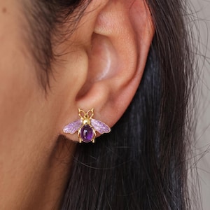 Può includere: Orecchini a lobo a forma di ape color oro con ali in smalto viola e una gemma viola centrale. Gli orecchini sono progettati per assomigliare a un'ape, con dettagli intricati sulle ali e sul corpo.
