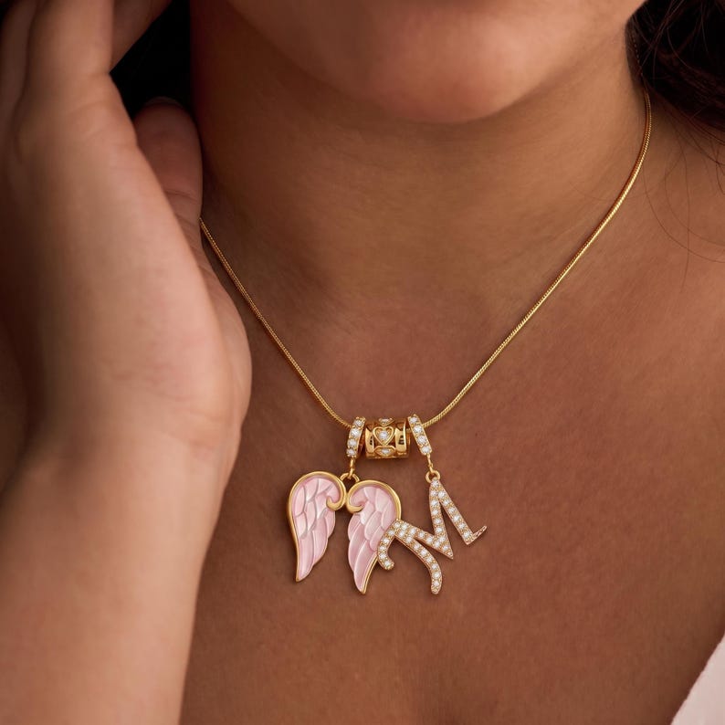 Puede incluir: Un collar dorado con una cadena fina. Presenta un dije con alas de &aacute;ngel de esmalte rosa y una letra "M" adornada con peque&ntilde;os cristales transparentes. Tambi&eacute;n hay una cuenta decorativa con un dise&ntilde;o de coraz&oacute;n.