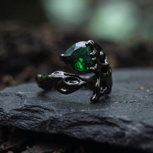 May include: A black metal ring featuring an open, textured band that resembles organic, gnarled branches. The ring is set with a single, faceted, pear-shaped emerald green gemstone held in place by dark, claw-like prongs on one side.