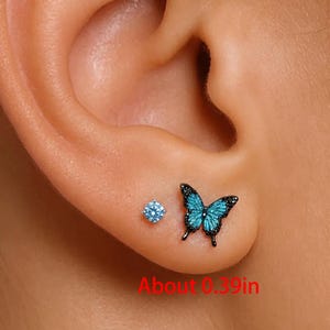 May include: An ear with two earrings is shown. One is a small, round, light blue stud. The other is a butterfly-shaped earring with turquoise wings and black accents. The butterfly earring is about 1 cm in size.