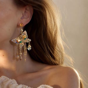 May include: A gold-toned dangle earring featuring a butterfly motif encrusted with amber, green, and clear crystals. The butterfly is accented with dangling gold chains and faceted crystal drops, suspended from a single amber-colored crystal stud.