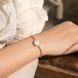 May include: A gold-colored bracelet with a round, white, crystal-like stone set in the center. The bracelet is delicate and thin, with a simple design. The stone has a textured appearance.