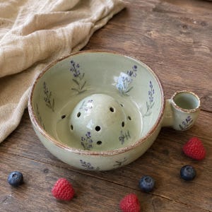 Pode incluir: Uma taça de cerâmica verde-clara para frutos silvestres com uma cúpula central com orifícios de drenagem e uma pequena pega lateral. A taça é decorada com ramos de lavanda roxos pintados à mão e tem um acabamento salpicado com rebordo castanho.