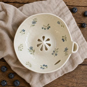 May include: A cream-colored ceramic berry bowl with a speckled finish, featuring a central drainage hole pattern shaped like a flower and small blue floral and berry illustrations scattered across the interior surface, including a side handle.