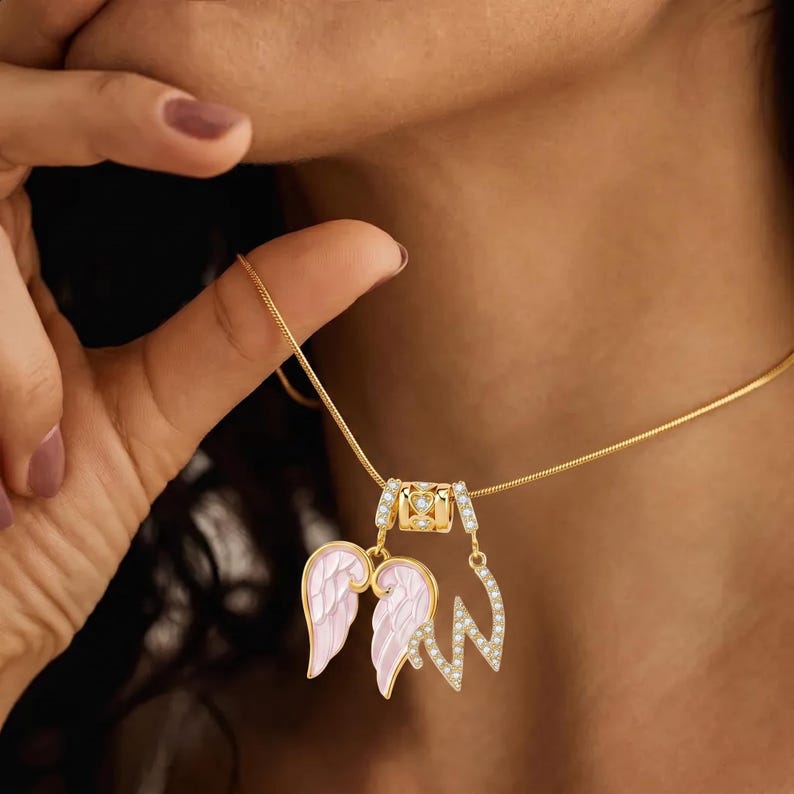 Puede incluir: Un collar dorado con un colgante que presenta alas de &aacute;ngel rosas y una inicial con diamantes de imitaci&oacute;n. El collar tiene una cadena delicada y un peque&ntilde;o dije decorativo con piedras preciosas transparentes. Las alas tienen detalles blancos.