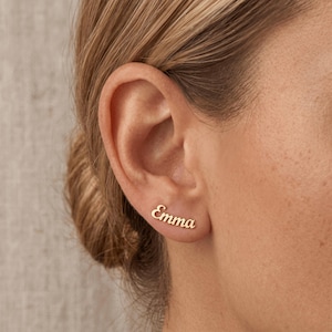 May include: A close-up of a person's ear with a gold earring spelling out the name "Emma" in a cursive font. The earring is a stud style, and the name is in a delicate, elegant script. The earring is set against the person's skin.
