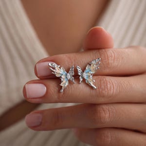 May include: A pair of silver butterfly earrings with light blue and cream-colored enamel wings. The earrings have small, clear gemstone accents and are displayed on a person's hand.