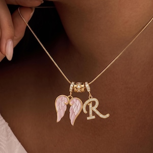 Puede incluir: Un collar dorado con tres colgantes. Los colgantes incluyen un par de alas de &aacute;ngel esmaltadas en rosa y blanco, una letra "R" con diamantes y una cuenta decorativa. El collar se muestra sobre un fondo oscuro.