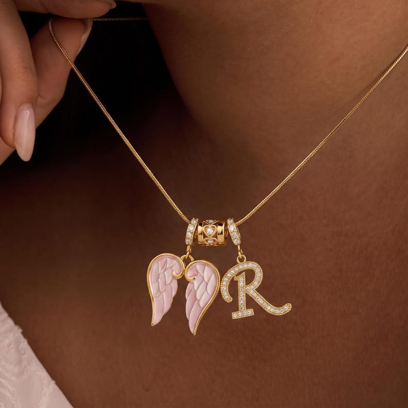 Puede incluir: Un collar dorado con tres colgantes. Los colgantes incluyen un par de alas de &aacute;ngel esmaltadas en rosa y blanco, una letra "R" con diamantes y una cuenta decorativa. El collar se muestra sobre un fondo oscuro.
