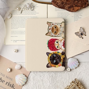 May include: A rectangular cream-coloured tablet case featuring a vertical illustration of three cats wearing colourful fruit-themed hoods. The top cat wears a strawberry hood, the middle cat wears a red berry hood, and the bottom cat wears a yellow floral hood.
