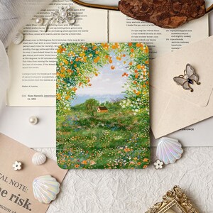 May include: A rectangular art print depicting a landscape with a cottage, a field of flowers, and a sky framed by orange blossoms. The print is surrounded by decorative items, including a butterfly brooch.