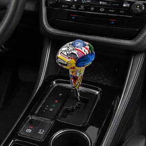 May include: A custom shift knob with a vibrant, multi-colored design featuring graffiti-style art and the Toyota logo. The knob is installed in a car's center console, with the gear shift indicator visible.