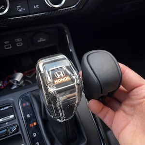 May include: A clear, faceted gear shift knob with the Honda logo in orange and white. A black, leather-like gear shift knob is held next to the clear knob. The background shows the interior of a car.