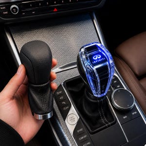 May include: A black leather gear shift knob next to a faceted, clear gear shift knob with blue LED lighting. The clear knob has the Infiniti logo illuminated. The car's console features buttons for various functions.