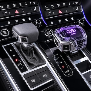 May include: A comparison of two car gear shifters installed in a centre console. One shifter is matte black with a leather boot, while the other is a transparent, faceted crystal-like material featuring the illuminated Audi logo in purple.