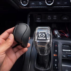 Może przedstawiać: Przezroczysta, fasetowana gałka dźwigni zmiany biegów z logo Mazda, obok czarnej skórzanej gałki dźwigni zmiany biegów. Dźwignia zmiany biegów znajduje się we wnętrzu samochodu z widocznymi pozycjami P, R, N, D i S.