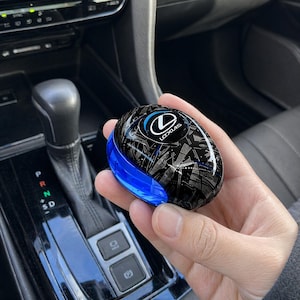 Peut inclure: Un étui de clé de voiture Lexus noir et argent avec un accent bleu. L'étui présente le logo Lexus en blanc et bleu. La clé est tenue dans une main, avec le levier de vitesse et la console d'une voiture en arrière-plan.