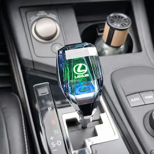 May include: A clear, faceted gear shift knob featuring a green and blue Lexus logo. The knob is in a car's console, near a wooden air freshener. The gear selector shows P, R, N, and S-D.