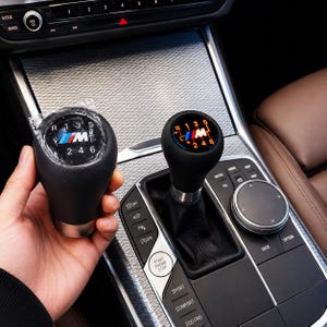 Puede incluir: Pomo de palanca de cambios de cuero negro con el logotipo BMW M y los números de marcha. El pomo se muestra junto a la palanca de cambios instalada en el interior de un coche. Los números de marcha están iluminados en naranja.