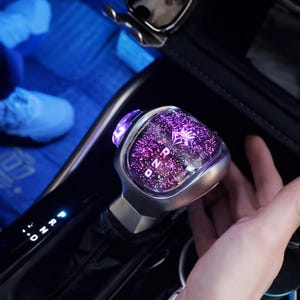 May include: A car gear shift knob with a purple and silver design. The top of the knob has a purple light and a pattern of small lights. The gear shift indicator shows "P R N D".