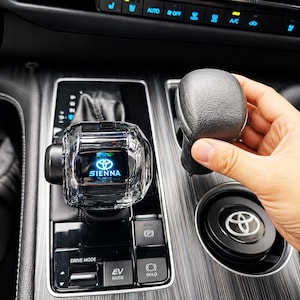 May include: A clear, faceted gear shift knob with the Toyota logo and "SIENNA" illuminated in blue. A black gear shift knob is held nearby. The image is taken inside a car, with the dashboard and console visible.
