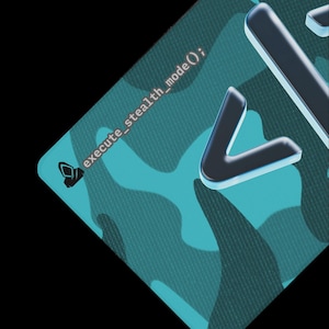 May include: A rectangular surface featuring a teal and dark green camouflage pattern. The text "execute_stealth_mode();" is printed in white, accompanied by a small icon of a hooded figure. Large, raised, metallic-edged characters are visible.