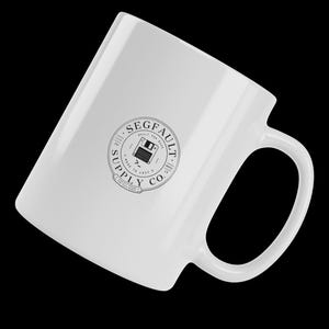 Può includere: Una tazza in ceramica bianca con un logo grafico circolare nero sul lato. Il logo mostra un'icona di un floppy disk al centro con il testo SEGFAULT SUPPLY CO. circostante, insieme alle frasi BUILT FOR BUGS e MADE TO LAST.