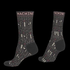 May include: A pair of dark gray crew socks featuring the word MACHINE in light pink at the top. The socks are decorated with a repeating pattern of white and pink technical symbols, including arrows, brackets, and progress bars on a dark background.