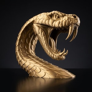 May include: A gold-coloured sculpture of a snake head with its mouth wide open, revealing sharp fangs and detailed scales. The serpent is depicted in a striking, aggressive pose with its neck curved, showcasing a metallic finish and texture.