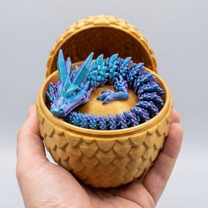 Può includere: Una piccola statuetta di drago articolata con scaglie blu e viola iridescenti, avvolta all'interno di un contenitore dorato a forma di uovo testurizzato. Il drago presenta corna cristalline e un corpo segmentato all'interno del guscio.