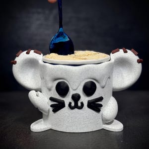 Può includere: Un contenitore in ceramica bianco a forma di topo con tratti del viso neri e accenti marroni sulle orecchie. Un cucchiaio blu è inserito nel contenitore, che è riempito con una sostanza beige. Il contenitore è su una superficie scura.