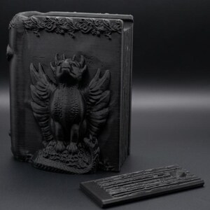 Könnte beinhalten: Ein mattes, schwarzes, rechteckiges und aufrecht stehendes buchförmiges Objekt mit einem Relief eines geflügelten Wasserspeiers auf dem Vorderdeckel. Der Deckel zeigt dekorative Schnörkel am oberen Rand. Ein separates, dünnes, rechteckiges schwarzes Teil liegt flach daneben.
